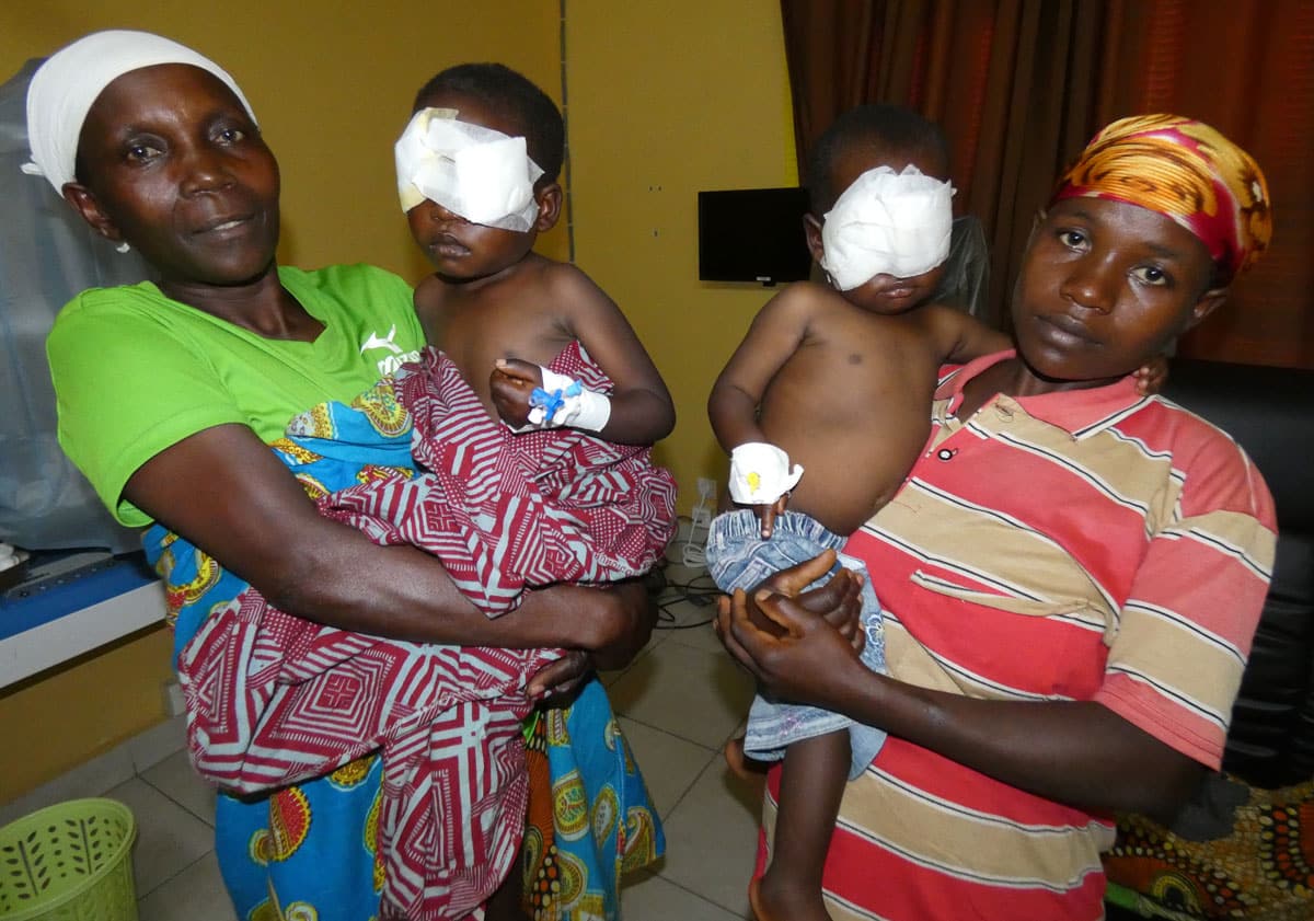 Wanawake wawili wawashikilia watoto wadogo wakiwa na bandeji juu ya macho yao