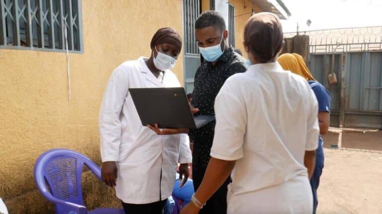 A man holding a laptop speaks to two medical professionals outdoors