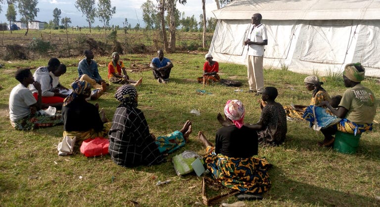 A group of adults in Burundi sit on the ground and listen to a standing speaker