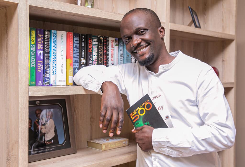An African man poses with a book by a bookshelf