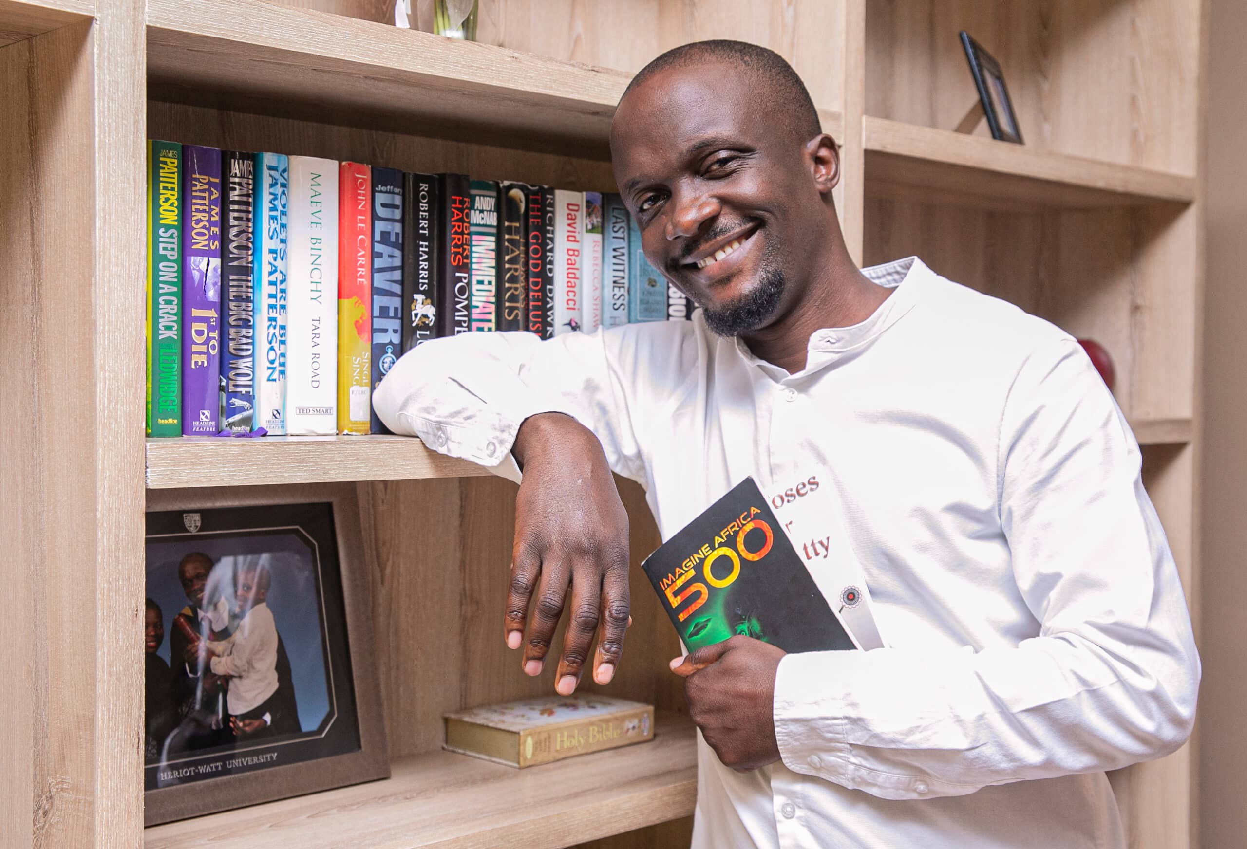 An African man poses with a book by a bookshelf