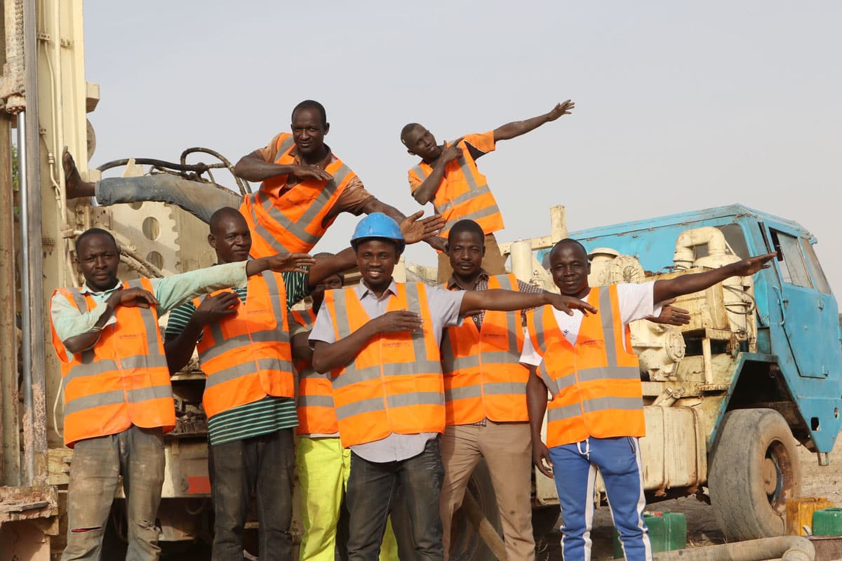 Seven African men in hard hats and vests stand by a water truck