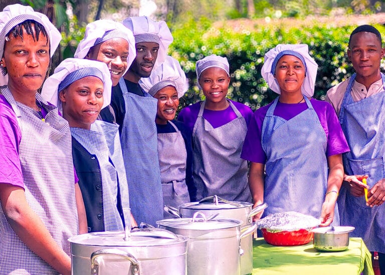 Eight Tanzanian students wearing purple and hairnets smile by a table of food